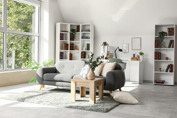 Interior of light living room with grey sofa, tables and bookshelves