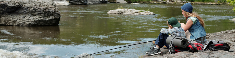 Woman sitting beside boy fishing together on rocky riverbank, both facing water, fishing rod...