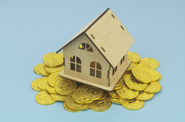 Wooden house on heap of cold coins on blue background.