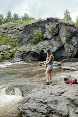 Woman standing on rocky riverbank fishing with rod near flowing water, wearing outdoor gear, backpack and camping equipment resting on rocks nearby