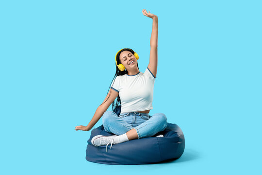 Beautiful young happy African-American woman in headphones sitting on beanbag chair against blue background