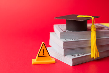 Yellow warning sign, graduation hat mortarboard, heap US dollar banknotes on red background....
