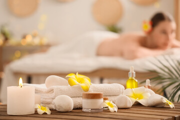 Towels with spa stones, candles, plumeria flowers, cosmetic products at table and young woman lying...