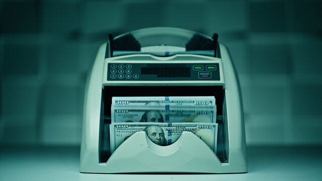 Machine rapidly processes bills beneath glowing lighting indoors. Dollars cash