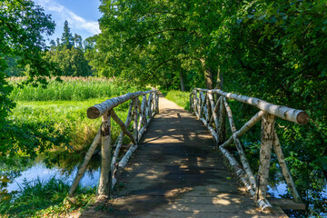 Wooden bridge with birch branch railing over stream in park, surrounded by green trees and grass, summer natural landscape