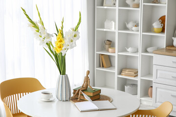 Vase with gladiolus flowers, books and cup on dining table in kitchen