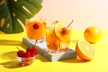 Glasses of tasty Old Fashioned cocktail with cherries and monstera leaf on yellow table against beige background