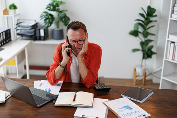 Stressed mature entrepreneur receiving bad news while talking on mobile phone and sitting at desk...