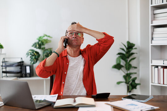 Worried businessman receiving distressing news during a phone call, pressing his forehead with his hand in a contemporary office filled with paperwork and technology