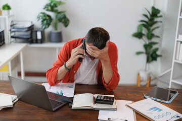Businessman receiving bad news while talking on the phone sitting at his desk in the office, he's...