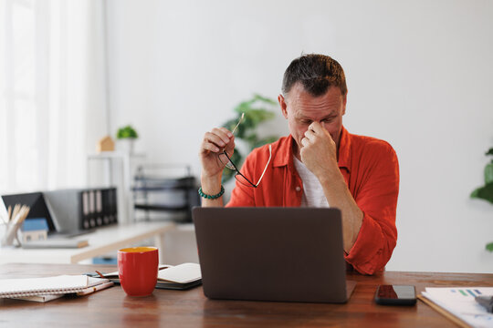 Stressed mature entrepreneur taking off glasses and rubbing his eyes while working on laptop at home, suffering from dry eye syndrome or eye strain after long computer work