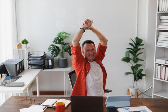 Mature entrepreneur stretching his arms while sitting at his desk in a modern home office, savoring a well-deserved break and relishing the moment of relaxation and comfort