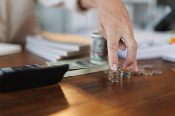 Accountant is stacking coins on a wooden desk, with dollar banknotes, a calculator and documents in the blurred background, depicting financial management and accounting concepts