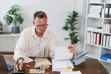 Focused mature entrepreneur sitting at desk and examining financial documents, calculating expenses...