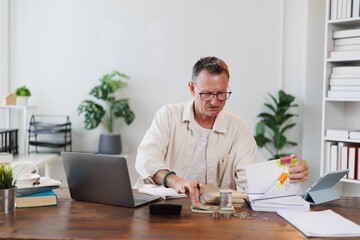 Focused mature entrepreneur calculating home budget and managing domestic finances, sitting at desk...