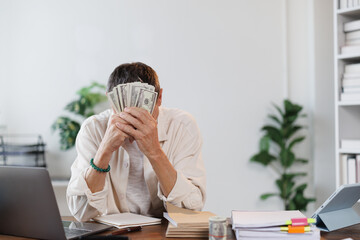 Businessman covering his face with dollar banknotes while sitting at a desk in a modern office,...