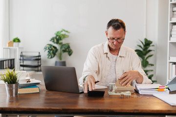 Focused mature accountant managing budget, calculating home finances and making notes while working with laptop and financial documents at home office