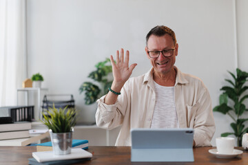 Happy teacher wearing glasses waving hand looking at digital tablet sitting at desk giving online...
