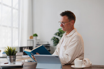 Obraz premium Senior businessman reading a book in his modern office, with a tablet next to him, taking advantage of natural light from a nearby window, creating a calm and productive work environment