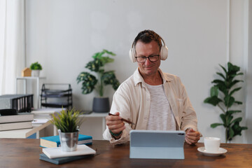 Mature entrepreneur wearing headphones and using a stylus pen participates in an online meeting on...
