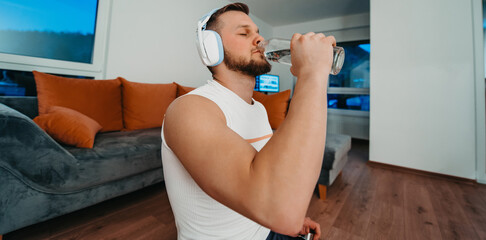 Close up of athletic man drinking water from bottle after training at home, headphones and...