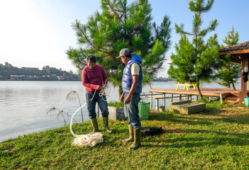 Two Men With Fishing Gear, Prepare Large White Net Beside Lake With Lush Trees