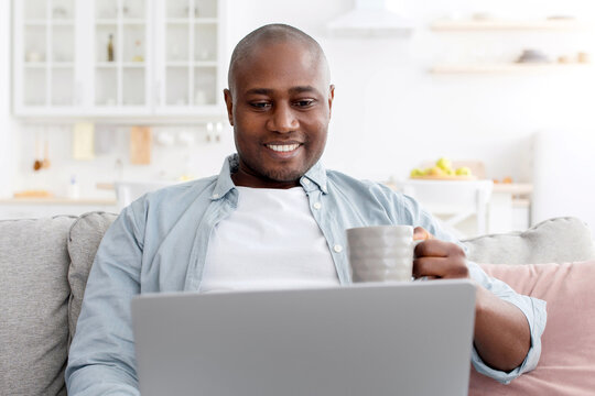 Reading morning news. Happy african american man using laptop and drinking hot coffee, smiling and looking at computer screen while resting on sofa at home, enjoying weekend