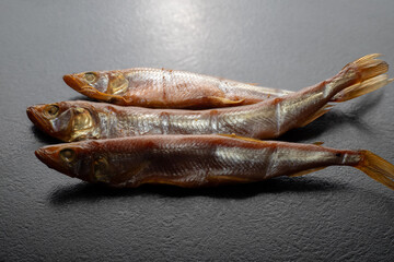 Close-up: cold hot smoked smelt in plastic container on black table on wooden board, top view. Smoked fish smelt. Sale of smoked smelt fish.