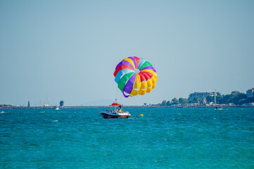 Two people in yellow life jackets are lifted into the sky by a vibrant parasail, soaring above the sea with a coastal town in the distance.