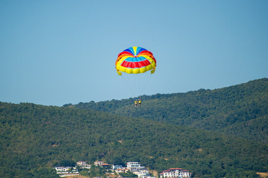 A parasailer floats above a scenic town and forested hillside, tethered to a vivid red, yellow, and blue parachute in Bu.garia