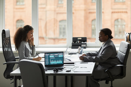 Two women, one Black and one biracial, working at office desk with laptops and documents, biracial woman talking on phone while Black woman reviewing paperwork