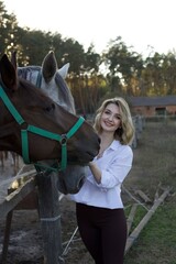 A young woman stands beside two horses, gently touching one while smiling. The setting is a...