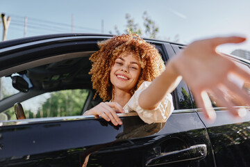 Smiling woman with curly hair reaching out hand from car window on sunny day, expressing joy and...