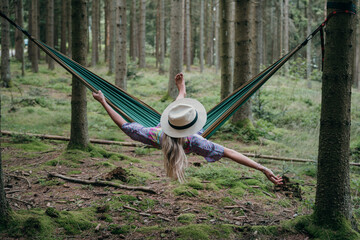 Traveler with a hat relaxes in a forest hammock. Peaceful moment in nature, viewed from behind.