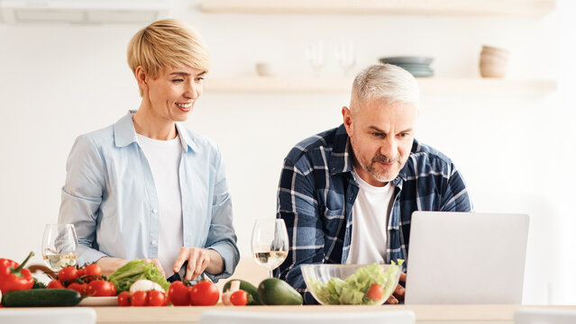 Search for recipe on Internet, cook food and communicate with blog followers. Concentrated middle aged man work on laptop, woman preparing dish, on table with vegetables in modern kitchen interior