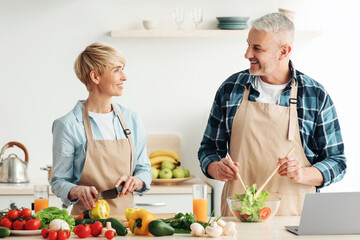 Free time together, preparing lunch for family and having fun. Happy mature husband and wife make salad in kitchen interior with bright vegetables, glasses of wine and laptop on table, empty space