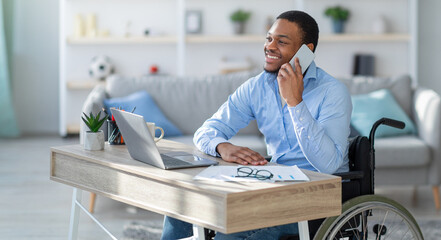 Joyful with disability black guy in wheelchair having phone conversation while working online on laptop from home. Young businessman talking with client on mobile phone