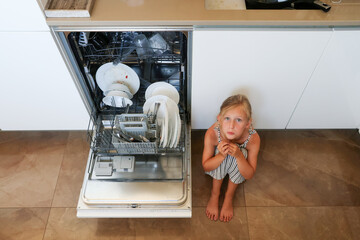 Sad child sitting on floor beside dishwasher in kitchen