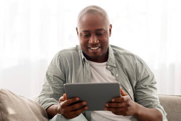 Mature black man spending time with digital tablet at home, reading news or browsing internet while relaxing on comfy couch in living room, enjoying weekend, free space