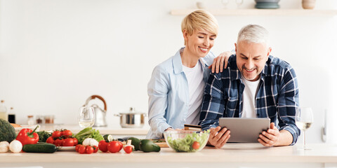 Happy senior couple use tablet, credit card, ordering food delivery in grocery in market doing shopping, buying online on website together at home in kitchen. Man and lady at table with vegetables