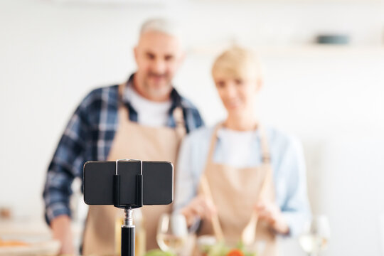 Video food blog, shooting and streaming. Smiling middle aged man and lady preparing salad and looking at smartphone webcam in kitchen interior with bright vegetables on table and glasses of wine