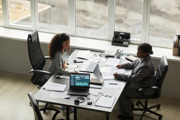 Two businesswomen, one Black and one biracial, working at office table, analyzing financial data on laptop and reviewing documents