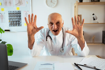 mature doctor with stethoscope works on a laptop looking at camera and showing up with fingers number ten. Senior therapist making video call, practitioner gp consulting patient online