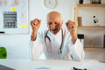 mature doctor with stethoscope works on a laptop looking at camera holding fists for good luck. Senior therapist making video call, practitioner gp consulting patient online