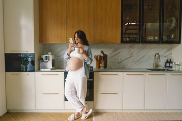Pregnant woman stands in a contemporary kitchen, focused on her smartphone. Natural light...