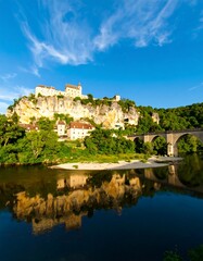 Fototapeta premium Picturesque village nestled on a cliffside overlooking a calm river with an arched bridge and its reflection
