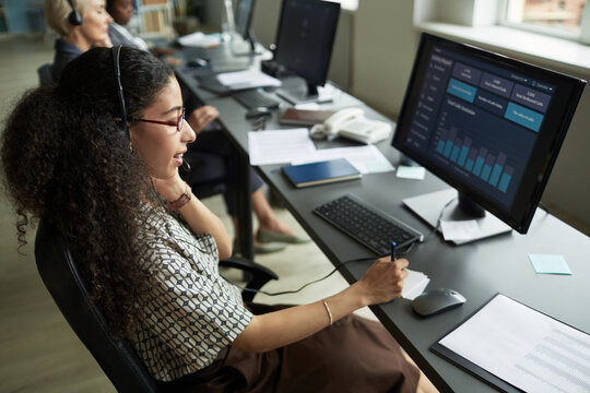 Young adult woman wearing headset working at computer desk analyzing data on monitor in modern office, colleagues sitting in background - Powered by Adobe