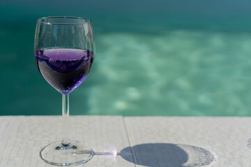 Purple drink in a wine glass on a pool edge a glass of purple liquid on a white tiled deck on swimming pool with a significant empty space on the right, resort lifestyle, luxury travel, party