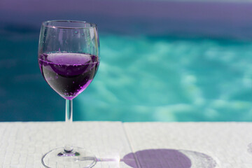 Purple drink in a wine glass on a pool edge a glass of purple liquid on a white tiled deck on swimming pool with a significant empty space on the right, resort lifestyle, luxury travel, party