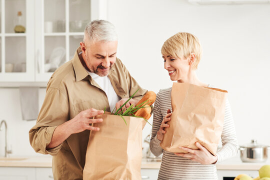 Buying products for family for diet and health care, online order and delivery. Smiling senior couple looks in paper eco bags, sorting out purchases in minimalist kitchen interior, free space